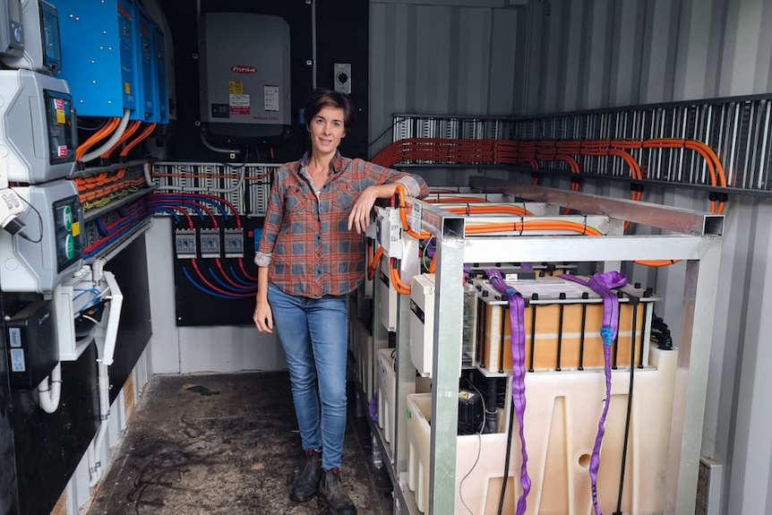 A woman with short dark hair, wearing a flannelette shirt, jeans and boots, stands in a shed full of solar batteries.