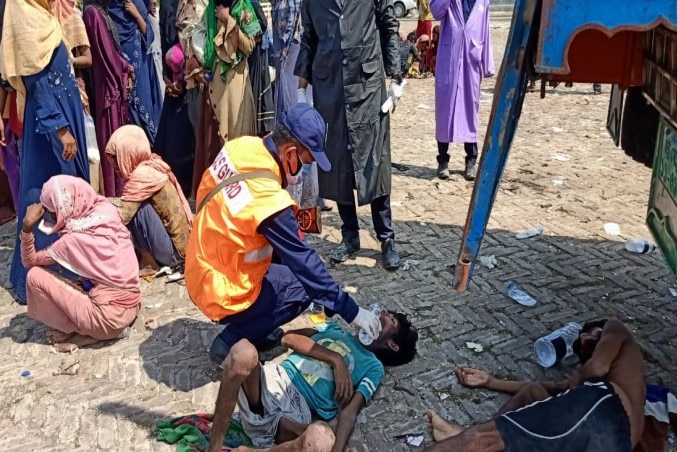 A coast guard in a high-vis vest assists a man lying on the ground with oxygen. Other refugees stand around them.