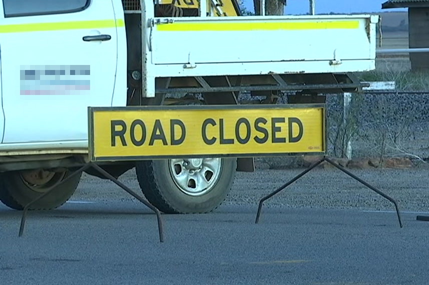 A yellow 'road closed' sign and a witches hat sit on the road near a ute.
