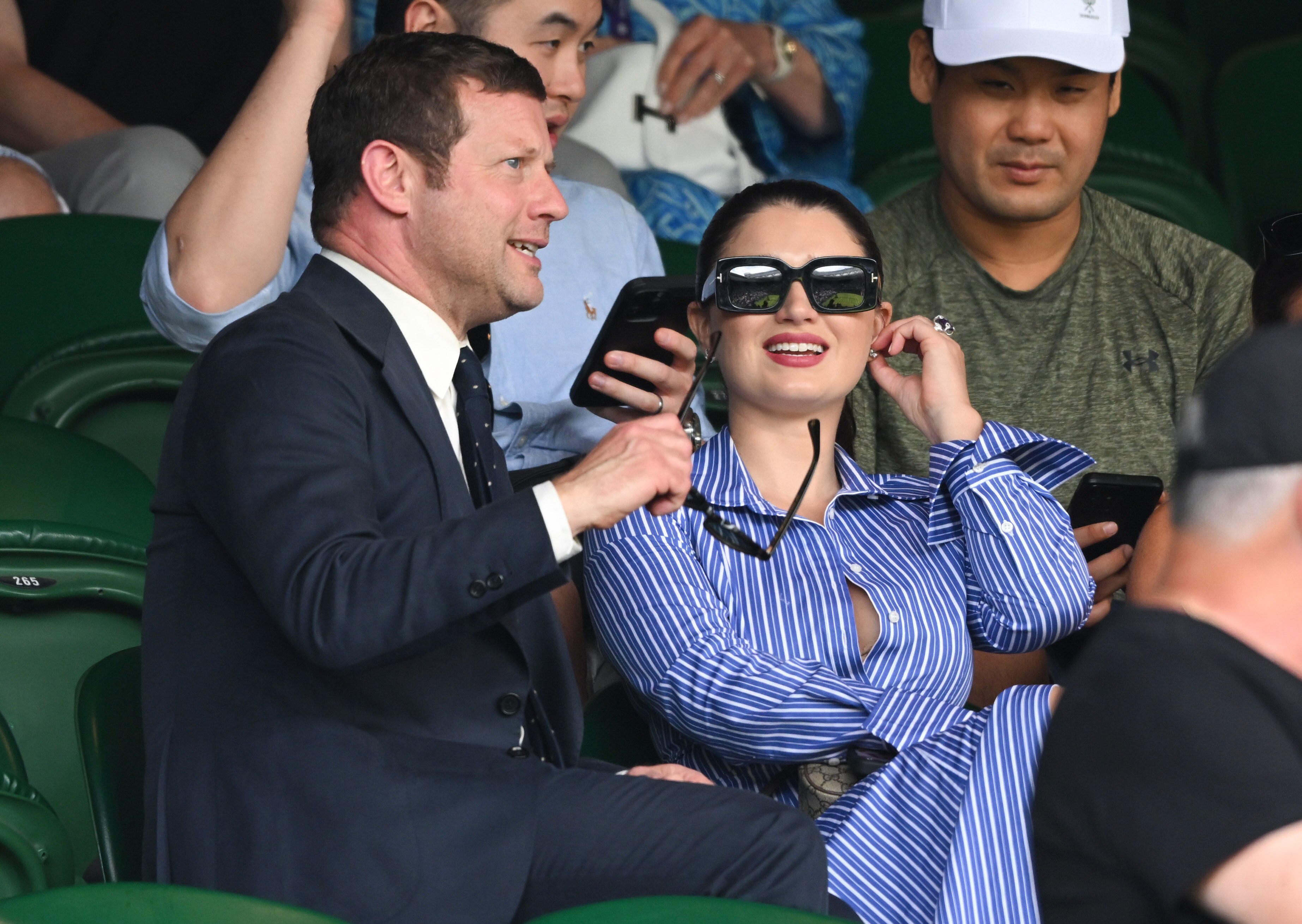 A woman in a blue striped shirt dress and dark sunglasses speaks wtih a man in a suit
