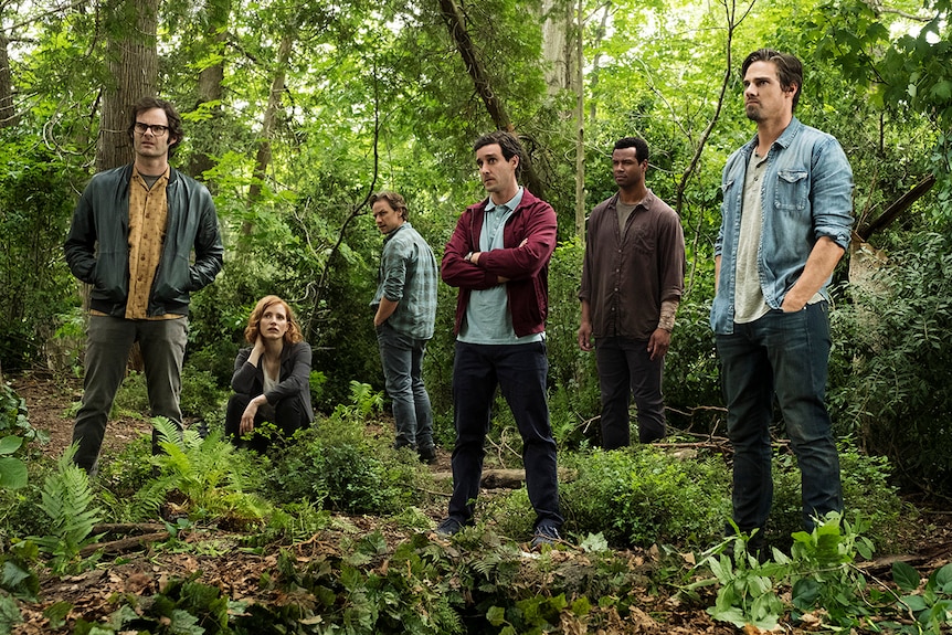 5 men stand and 1 woman crouches in green bushland looking umimpressed.