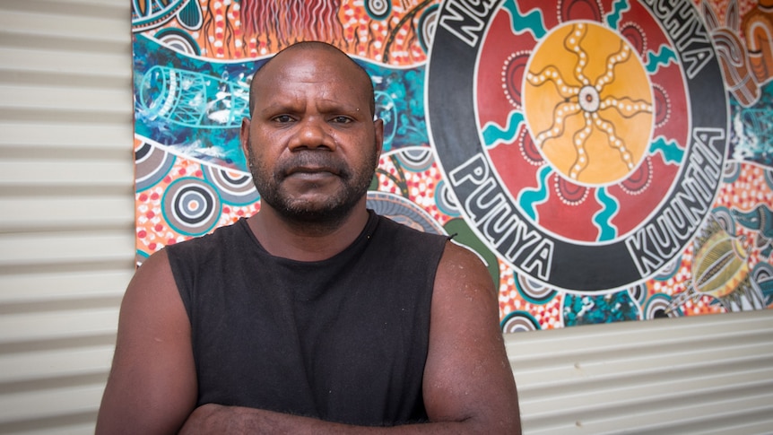 An Aboriginal man standing in front of a brightly coloured painted logo hung on a corrugated metal wall.
