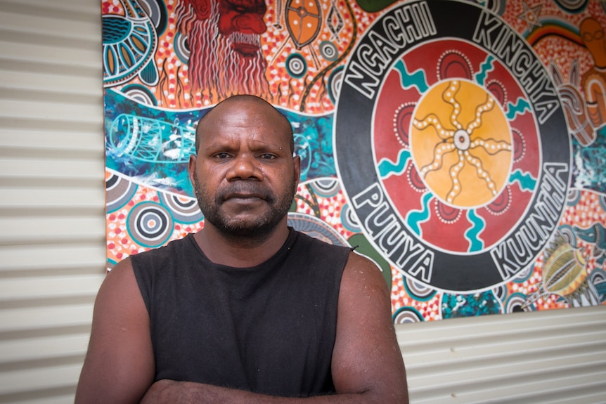 An Aboriginal man standing in front of a brightly coloured painted logo hung on a corrugated metal wall.