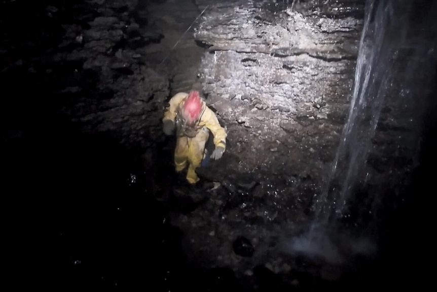 A person in yellow overalls and a red helmet in a cave with a waterfall 