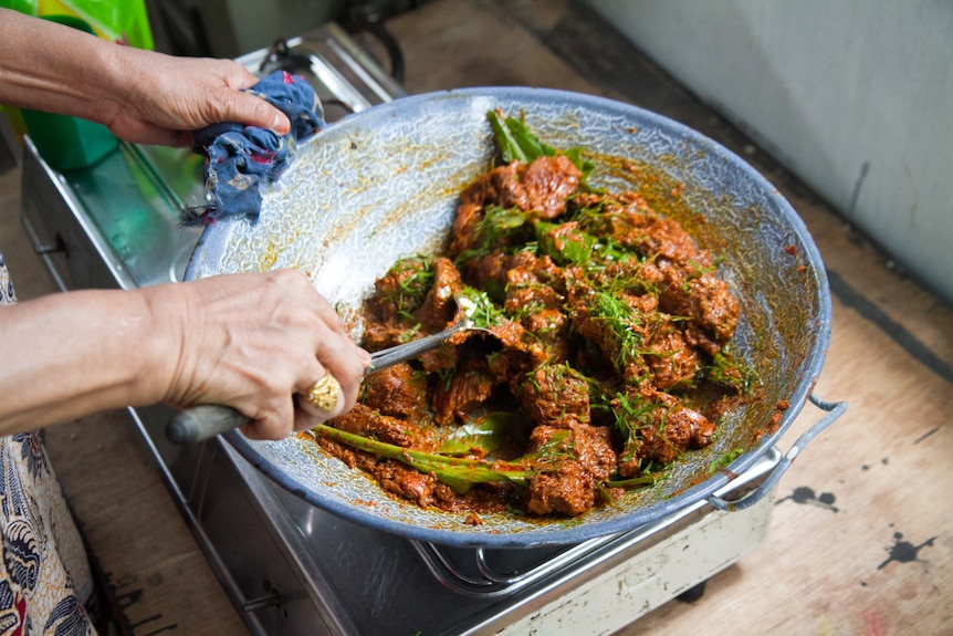 a hand stirs a wok of cooking beef rendang