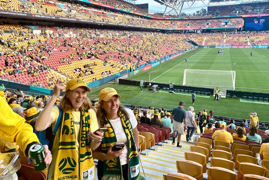 Two women, looking happy, wearing green and gold at a stadium.