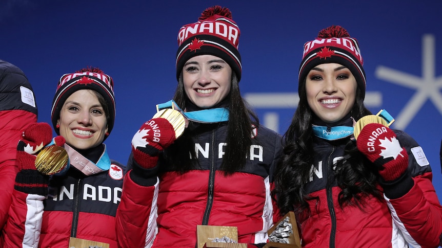 A trio of Canadian female figure skaters stand on a podium smiling wearing their Winter Olympic gold medals.