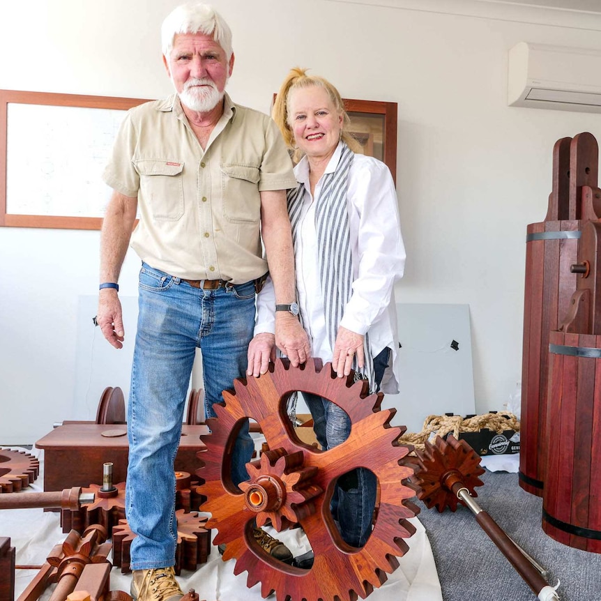 A man and woman standing up holding a up a wooden piece of a clock with more wooden clock pieces in the background.
