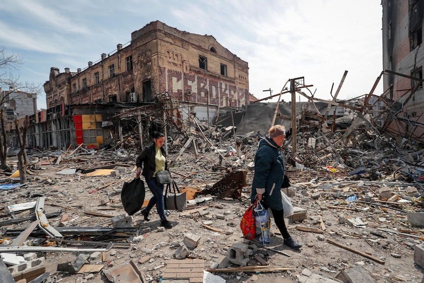 People walking with bags with ruins and debri in background