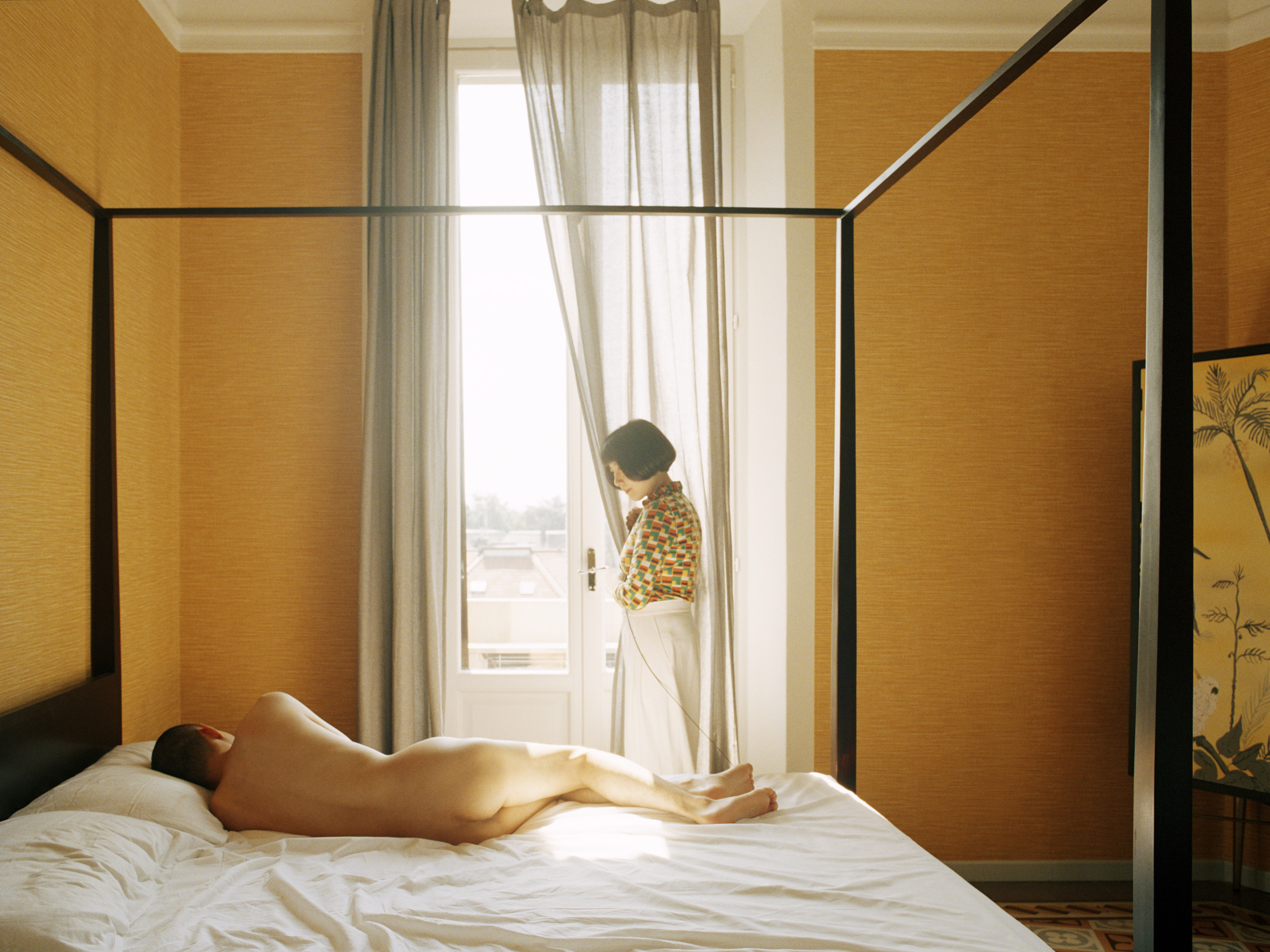A naked Japanese man lies on a four-poster bed in a yellow hotel room. A Chinese woman in a patterned shirt and white pants s