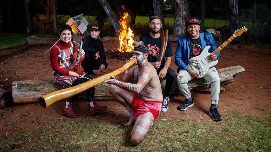 A group of instrumentalists sitting in front of a campfire, with a Yidaki and player in the center.