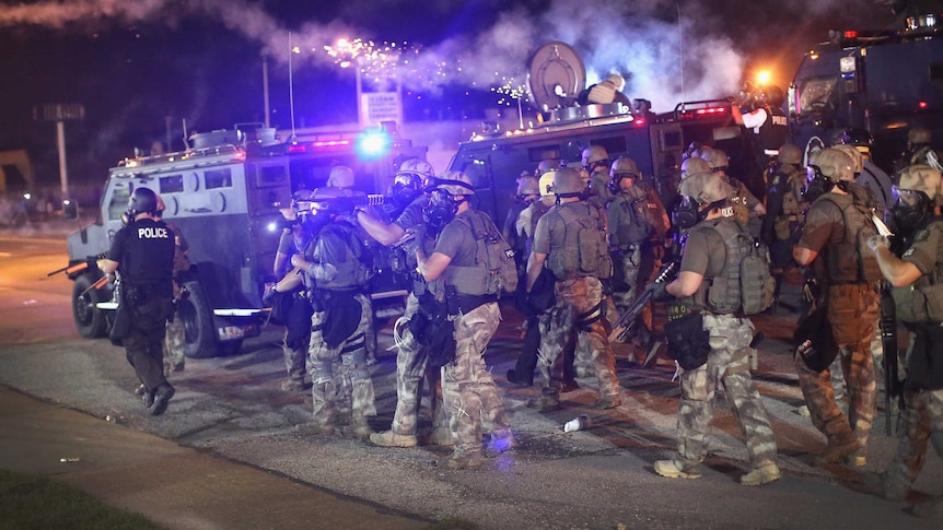 Police dressed in green fatigues walk alongside vehicles during protests.