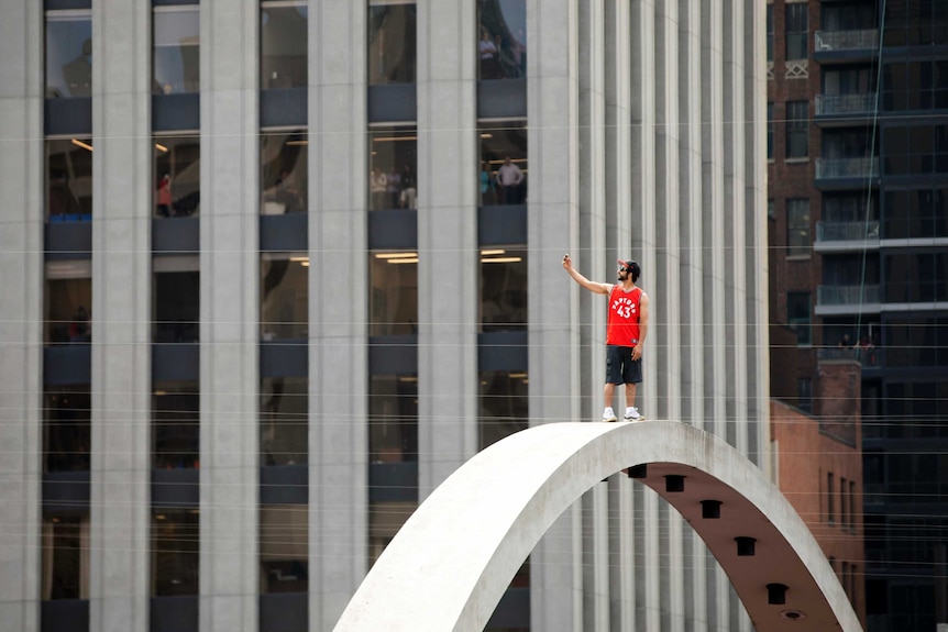 A man in a red basketball singlet stands on a large curved structure high off the ground as office workers watch from a building
