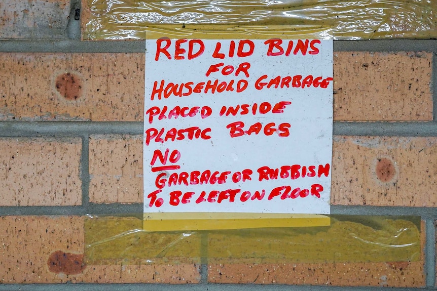 A sign stuck to a brick wall reads: "Red lid bins for household garbage placed inside plastic bags."