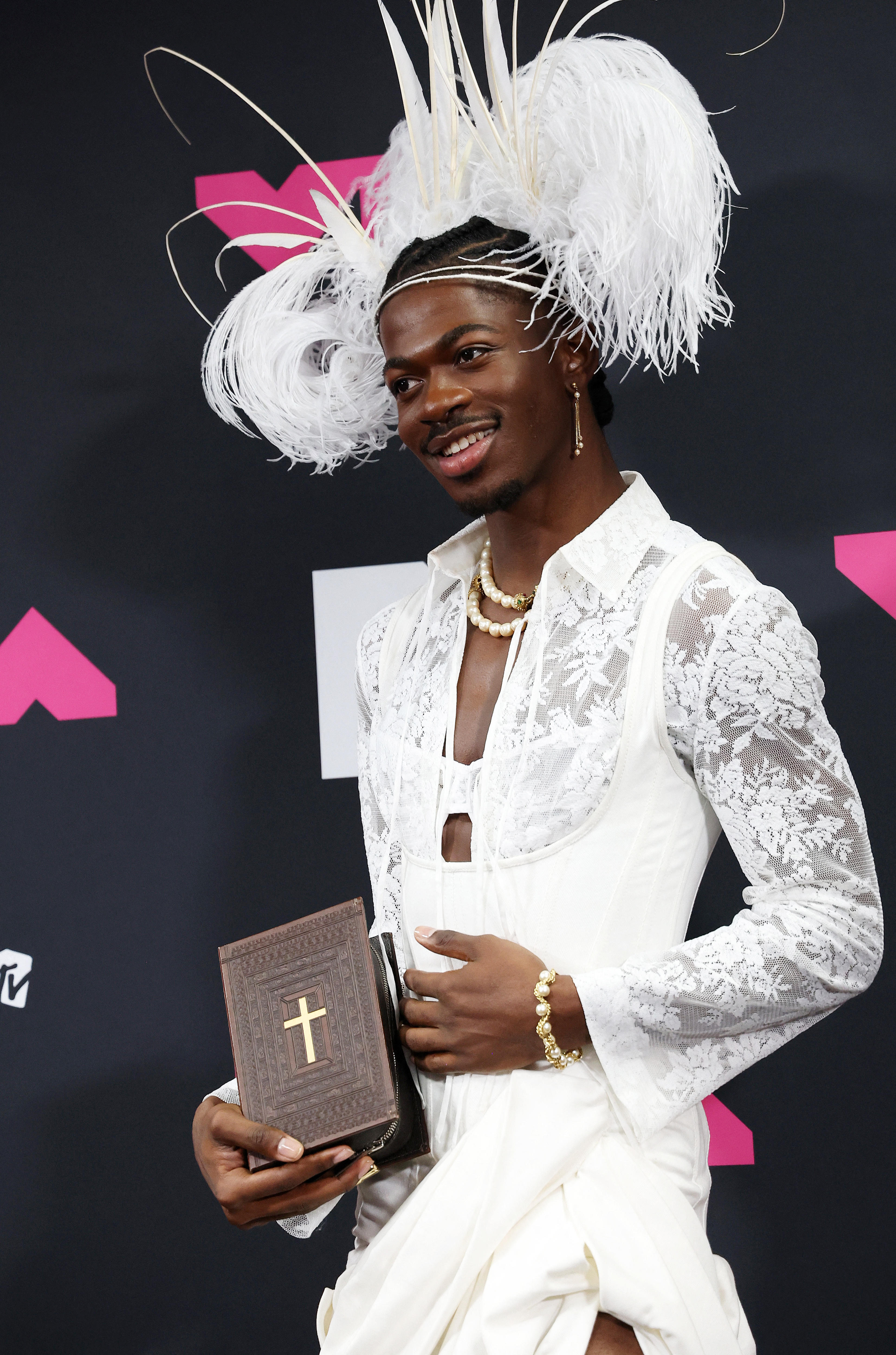 Lil Nas X poses wearing a white laced top, feathers on his head and a bible in his hands.