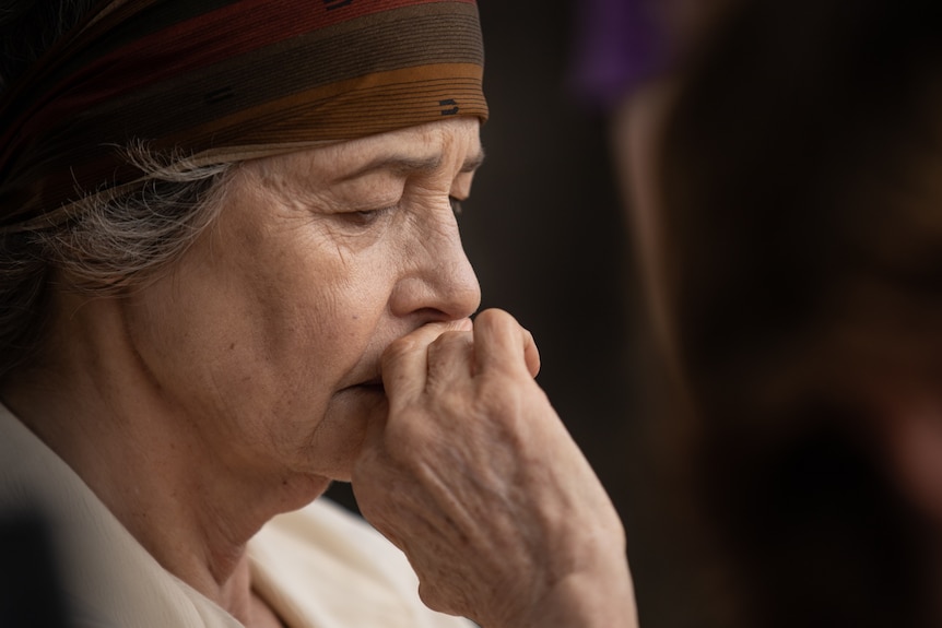 An old woman wearing a headscarf, looking down with her hand to her lips, seen close up from the side