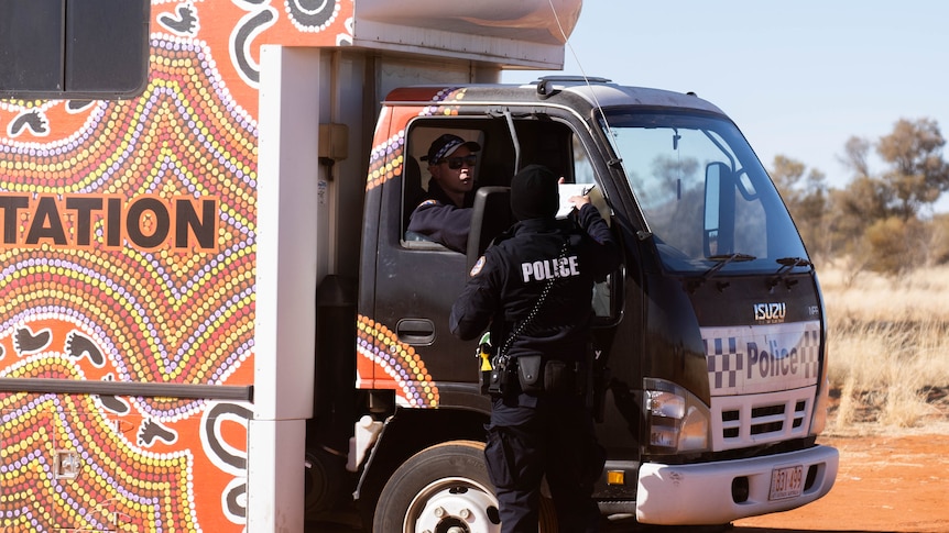 A police officer sitting in a police truck speaks out the window to another police officer, with desert in the background.