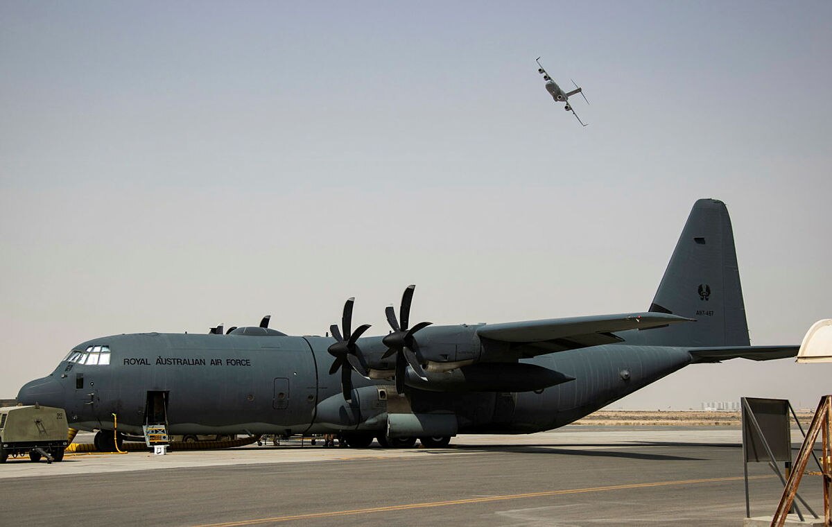 A large military plane rests on the tarmac, with a second plane visible in the air in the distance.