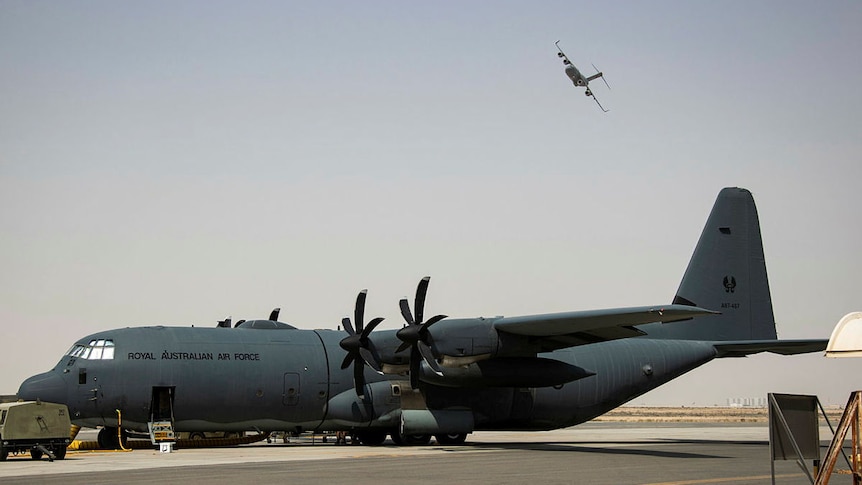 A large military plane rests on the tarmac, with a second plane visible in the air in the distance.