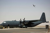 A large military plane rests on the tarmac, with a second plane visible in the air in the distance.