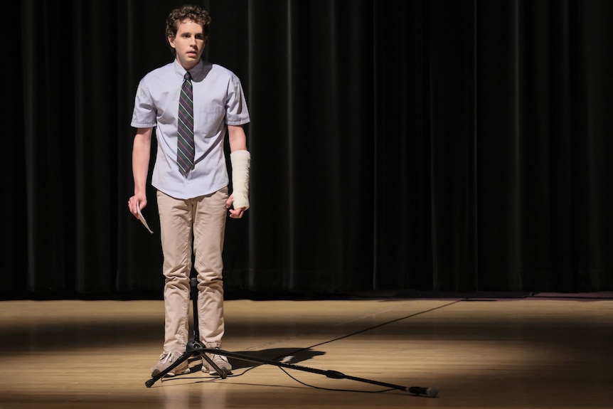 A young man with a broken arm in a cast looks worried, standing on a school stage, wearing a tie