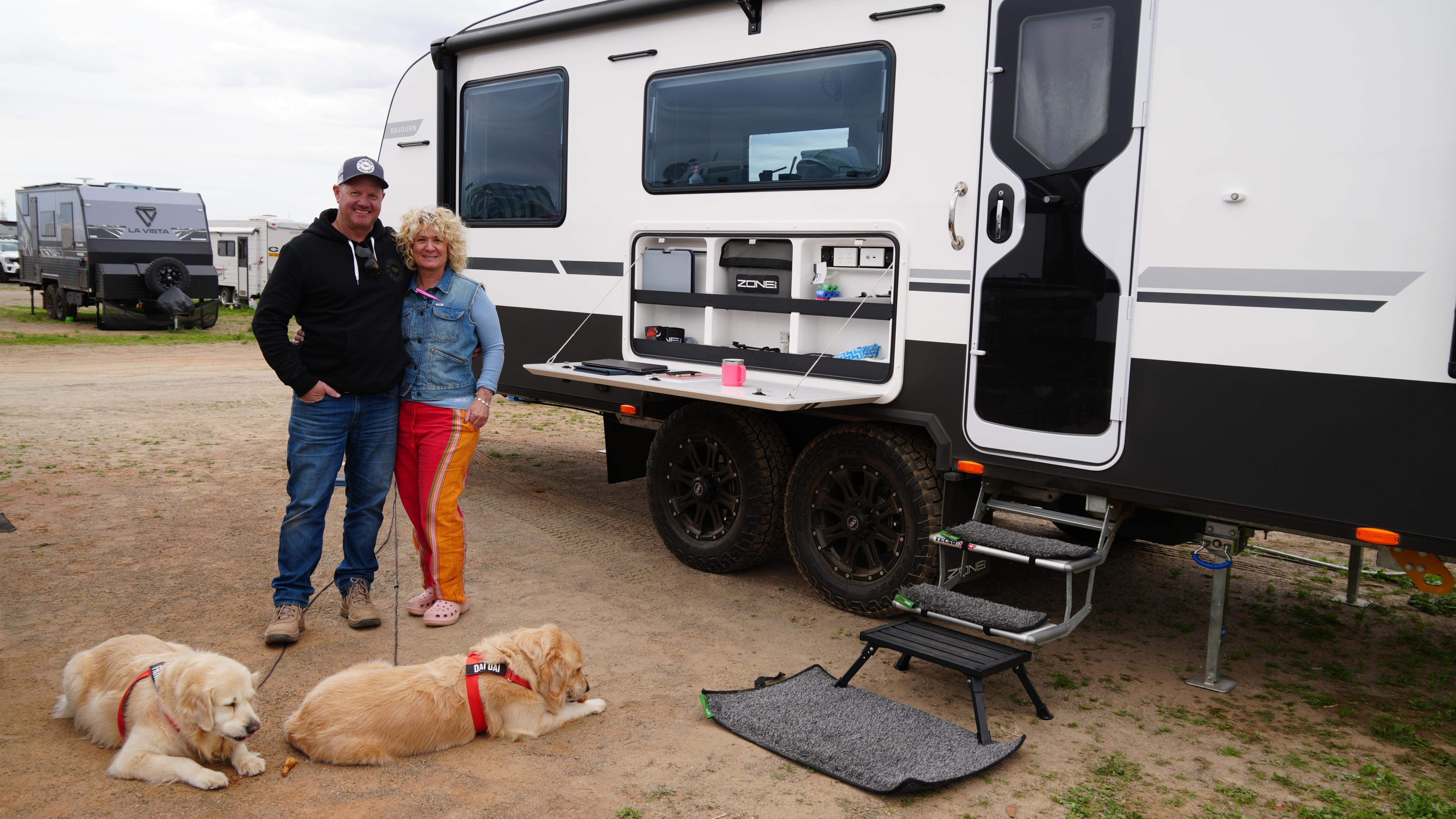 Man and woman stand next to white caravan with two golden retriever dogs in front of them on ground.