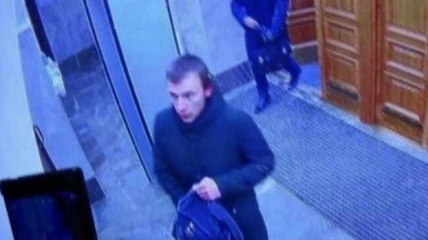 Still from video footage showing a teen boy with his hand inside a backpack in an office. He has a neutral expression.