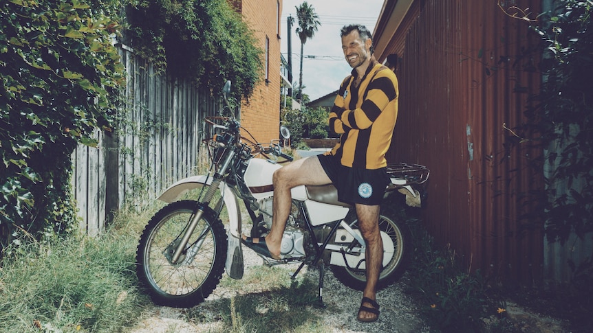 Felix Riebl next to a white motorbike with arms crossed, wearing a football jumper, grinning, wearing sandals