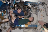 A male astronaut exercises in a confined area of the International Space Station. He is assisted by another astronaut.