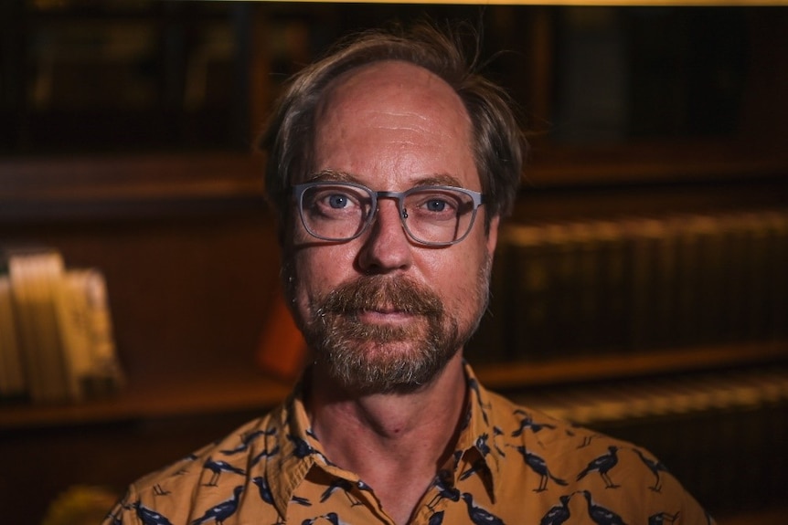 A fair-skinned, blond-haired man with a beard and glasses and an orange shirt.