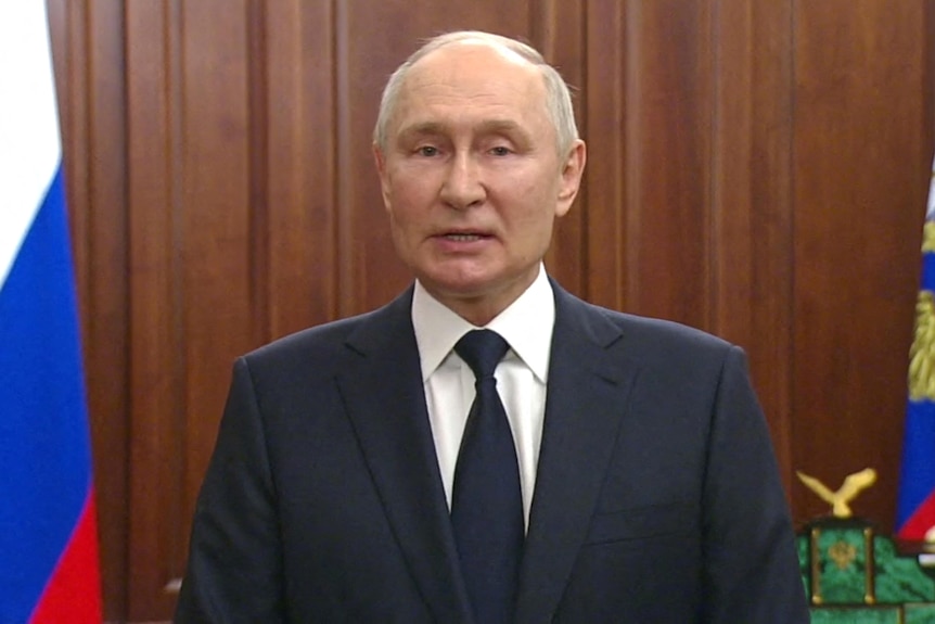 Vladimir Putin speaking in black suit and tie flanked by Russian flag.