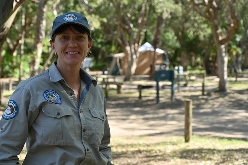 A woman in park ranger uniform stands smiling at a campsite with a tent in the background.