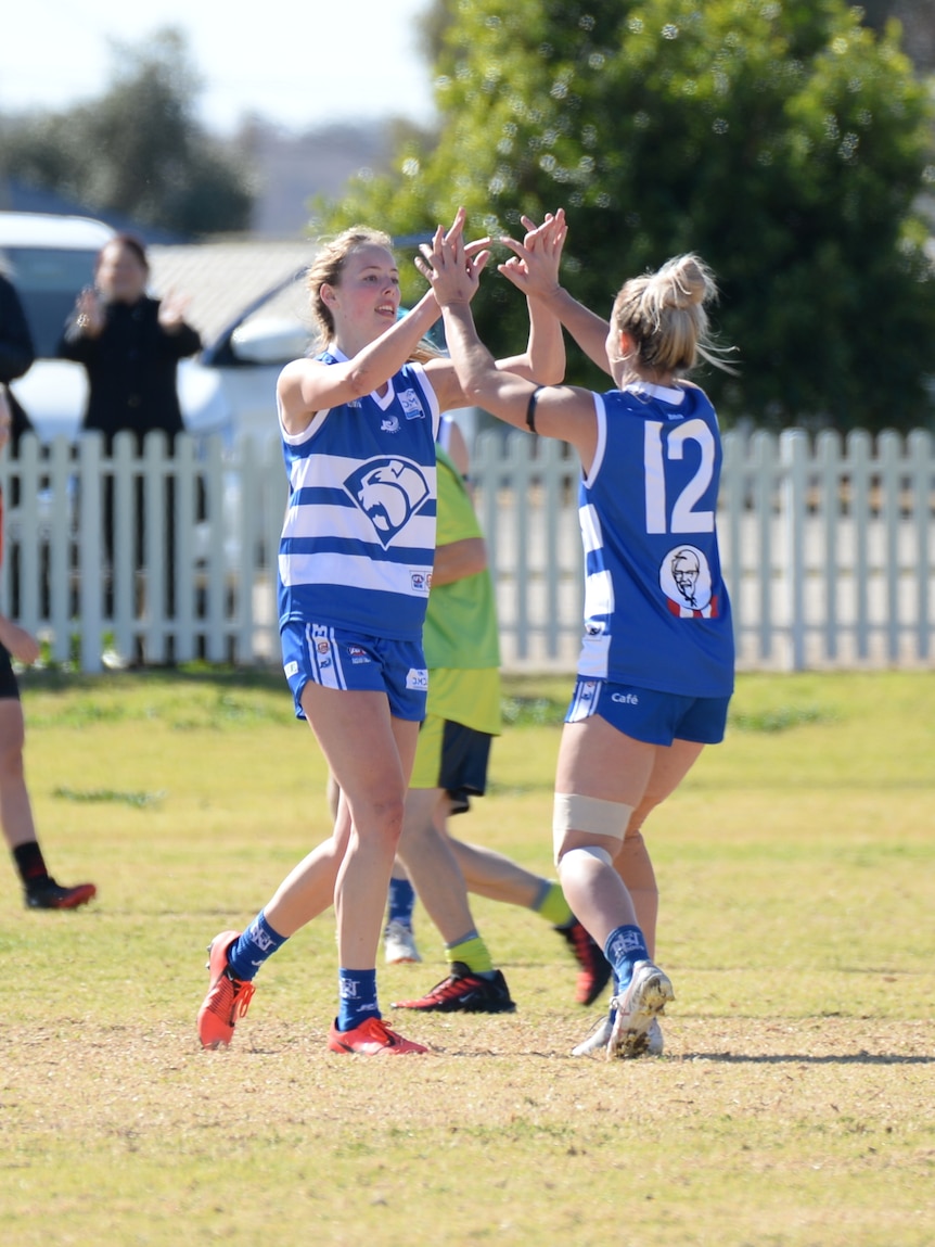 Deux femmes blondes portant un uniforme bleu avec un bouledogue célèbrent, tout en jouant à l'AFL