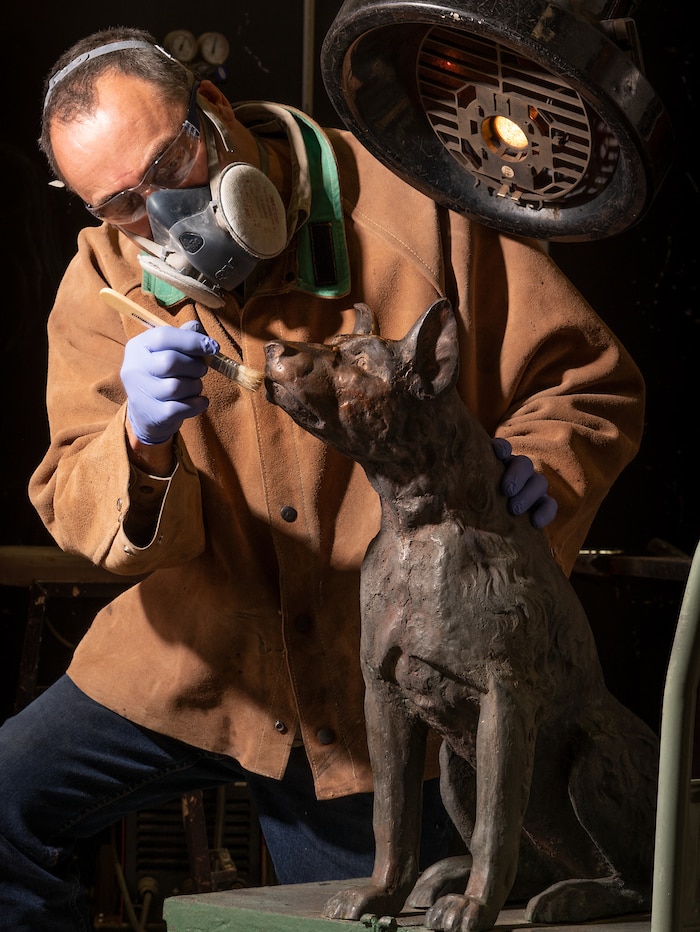 A man wearing a mask and gloves holds a paintbrush and works on a statue of a dog.