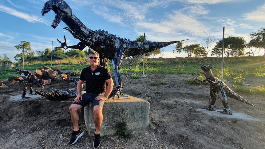 Steve Ross sits at the base of a large dinosaur sculpture smiling with other dinosaurs in the background.