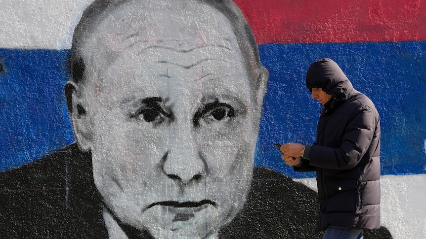 A man walks by a large mural of Vladimir Putin's face against a blue, red and white background. 