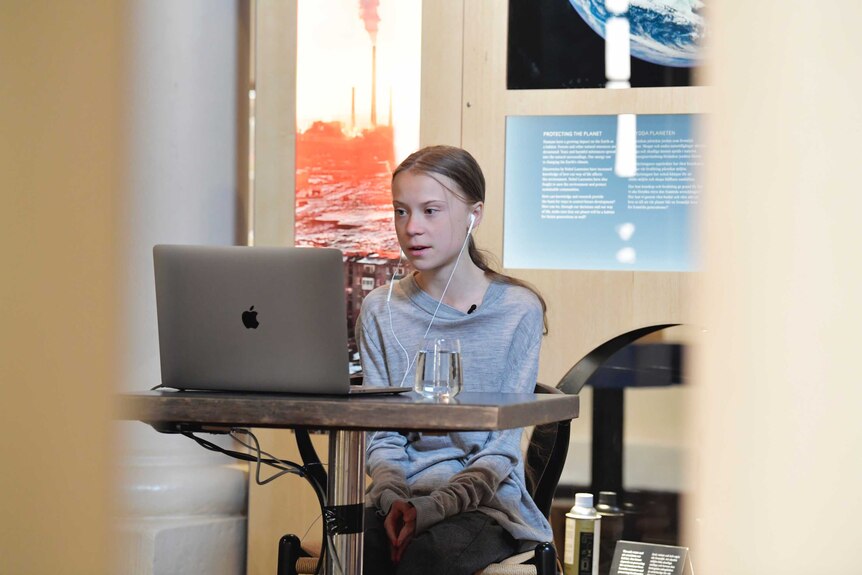 Greta Thunberg sitting at a table wearing a grey shirt and talking on a conference call on her laptop with her earphones in.