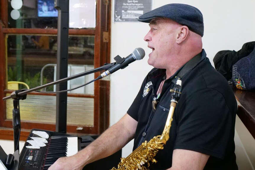 Mark McGurgan singing into microphone with a saxophone around his neck and playing piano. 