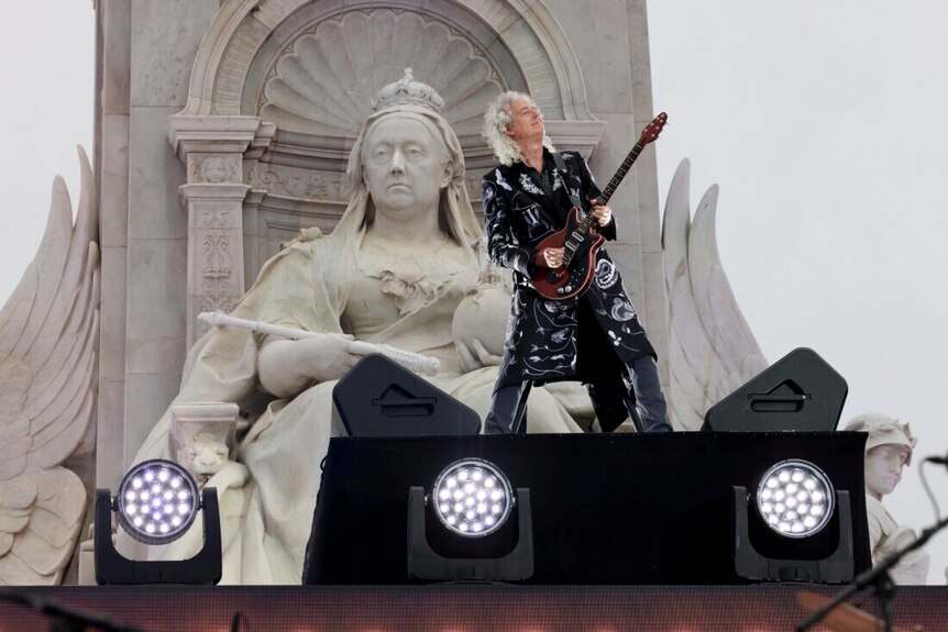 Брайан Мэй играет на гитаре перед большой статуей королевы Виктории в Букингемском дворце.  Выглядит счастливым. 
