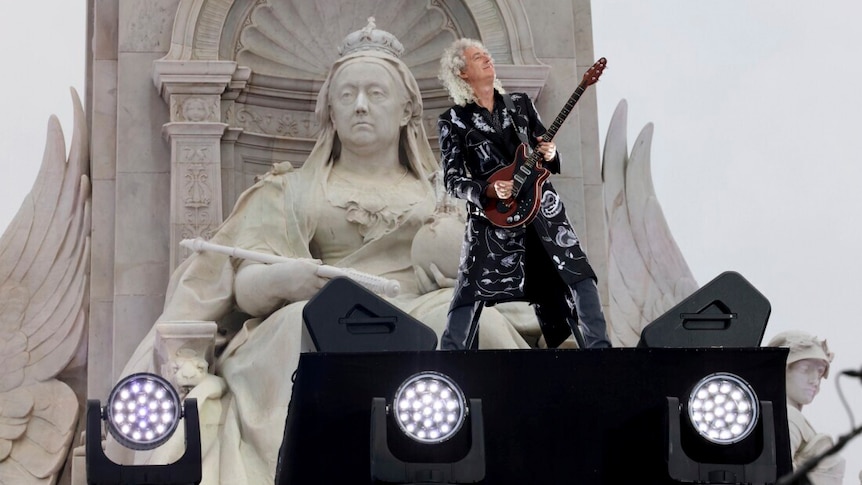 Brian May plays his guitar in front of a large statue of Queen Victoria at Buckingham Palace. He looks thrilled. 