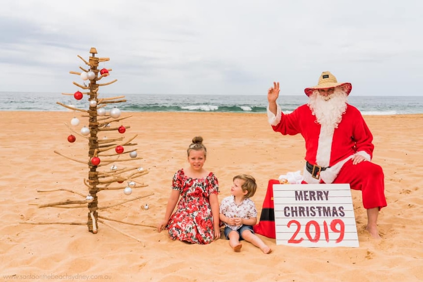 Santa sitting on the beach next to two young children, and a wooden Christmas tree.