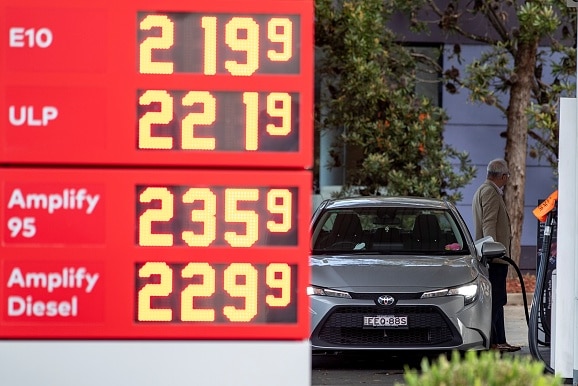 Ein rotes Schild listet die Benzinkosten auf, während ein Mann in der Ferne sein Auto betankt. 