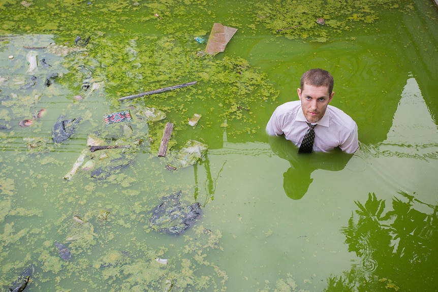 A man stands in green swampy water, wearing a business shirt and tie