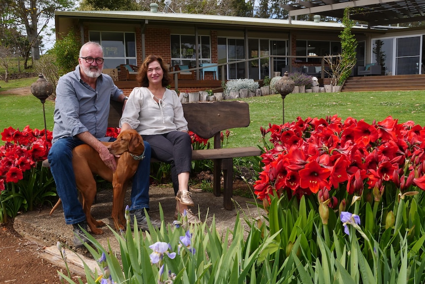 A man and woman sit on a bench in their backyard, with their dog, surrounded by red flowers.