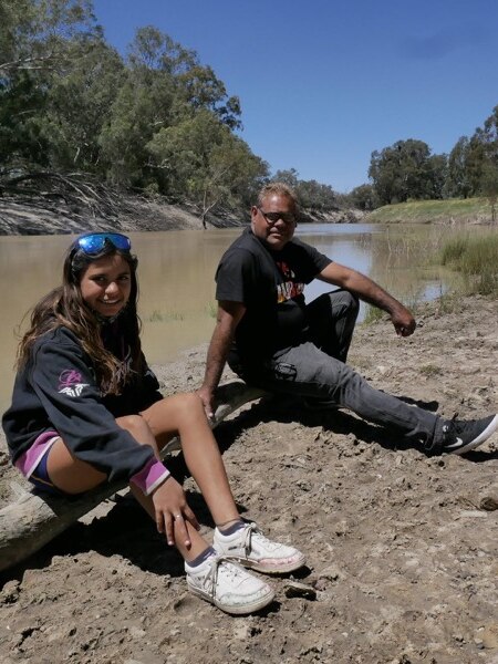 A girl wearing sunglasses and a man wearing a teeshirt and spectacles sit beside a river.