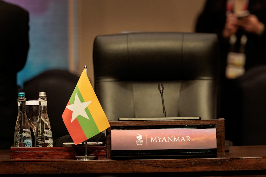 A flag with yellow, green and red stripes behind a white star sits on a desk next to MYANMAR name plate, by an empty chair