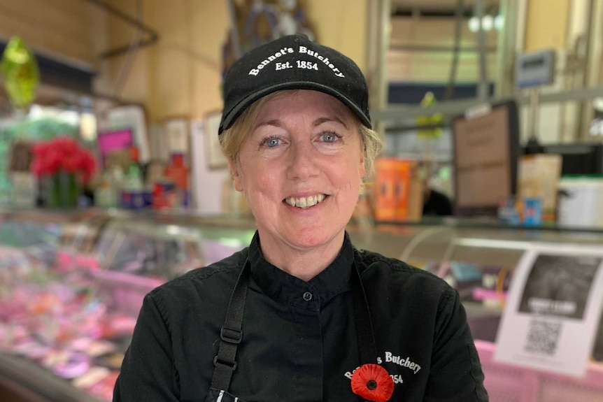 A smiling woman wearing a cap and apron stands inside a butcher shop.