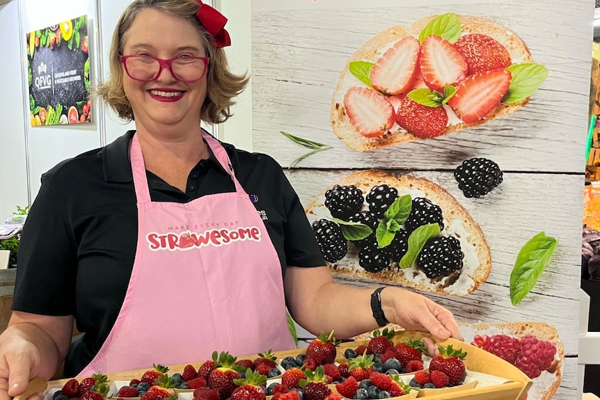 A middle-aged woman stands holding a platter of berry tarts, with a Berries Australia sign in the background.