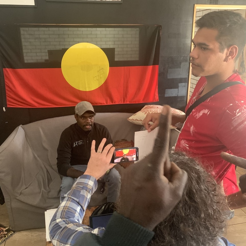 Someone films a man with an iPhone while he talks in front of a flag inside a building.