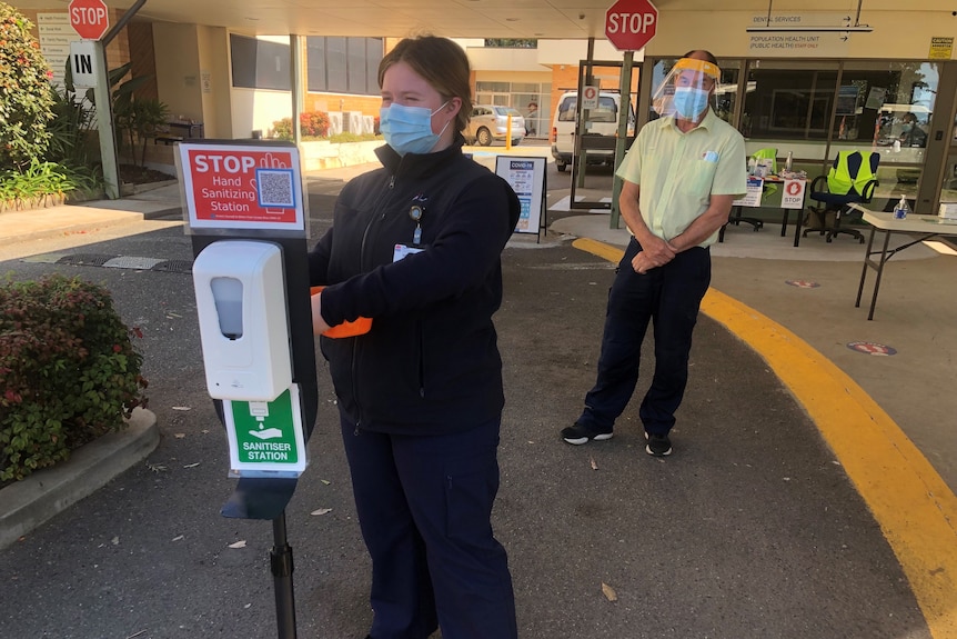 A woman with a mask on stands next to a hand sanitising station outside a local health office.